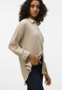 Light beige button-up shirt with textured fabric, long sleeves, and a curved hem is paired with dark denim jeans.