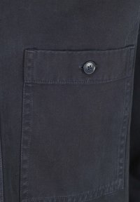 Zwart, gestructureerd stof toont een close-up van een hemdzak met een ronde, viergaatjesknop. Schone stiksels zichtbaar langs de randen.