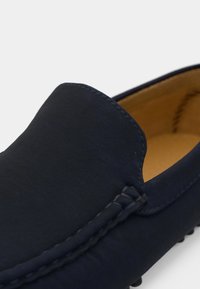 Gros plan d'un mocassin en daim bleu marine foncé avec des coutures visibles et une doublure intérieure couleur fauve.