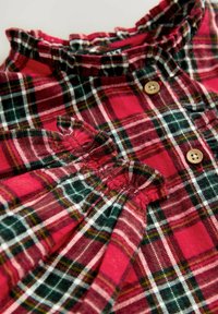 Chemise à carreaux rouge et verte avec un col froncé, des boutons en bois et des détails en tissu plissé. Matière en coton texturé.