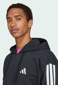 Sweatshirt com fecho éclair preto, com design de três riscas brancas nas mangas, material em mistura de algodão, capuz ajustável com cordão, logo discreto no peito.