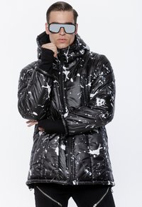 Mężczyzna w czarno-białej kurtce puchowej w plamy, z futurystycznymi lustrzanymi okularami, trzymając rękę przy brodzie, stojący na tle białym.