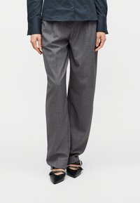 Άτομο που φοράει χαλαρά γκρι παντελόνια, μαύρες μυτερές γόβες με λουράκια στον αστράγαλο και σκούρο κουμπωτό πουκάμισο, στέκεται μπροστά σε απλό φόντο.