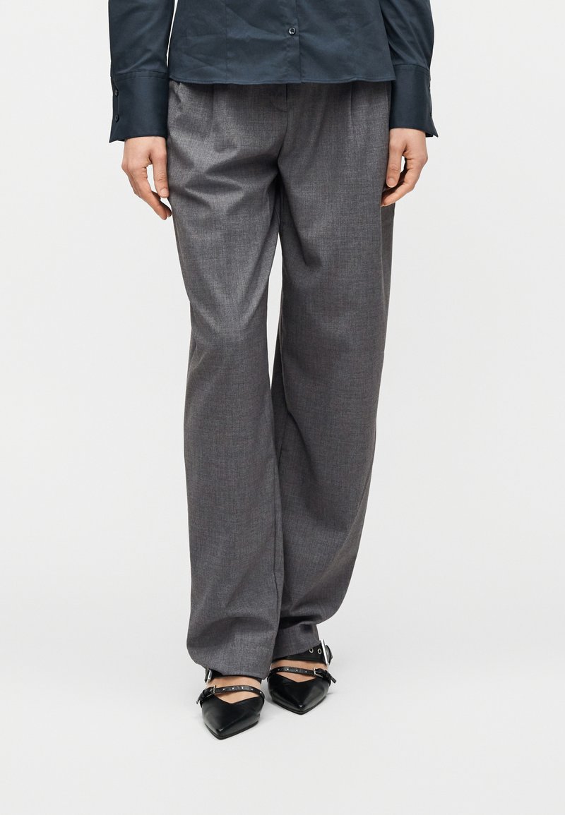 Άτομο που φοράει χαλαρά γκρι παντελόνια, μαύρες μυτερές γόβες με λουράκια στον αστράγαλο και σκούρο κουμπωτό πουκάμισο, στέκεται μπροστά σε απλό φόντο.