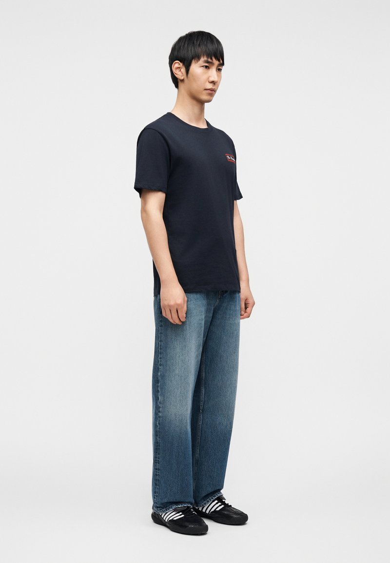 Navy blue cotton t-shirt with a small red logo, paired with loose-fitting light blue denim jeans and black shoes with white stripes.