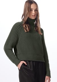 Maglione a collo alto in verde scuro, con un motivo testurizzato, polsini e orlo a coste. Indossato con pantaloni neri a vita alta.