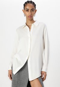 Camisa branca de botão com mangas longas, feita de um tecido leve. Apresenta um colarinho clássico e um corte solto, combinada com uma saia cinza.