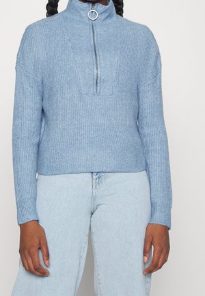 Maglione a maglia azzurro chiaro con collo alto e zip, caratterizzato da un motivo testurizzato e polsini a coste, abbinato a pantaloni di denim chiaro.