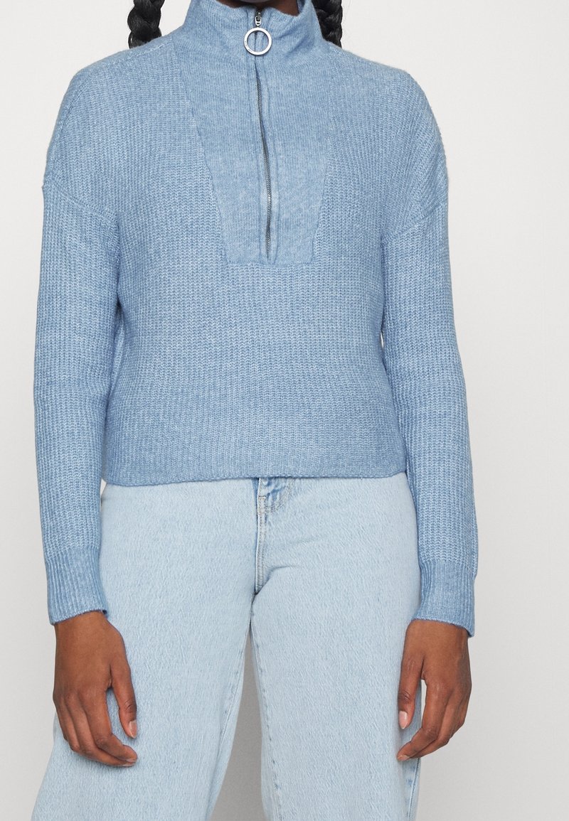 Ljusblå stickad tröja med hög krage och dragkedja, med ett strukturerat mönster och ribbade ärmslut, i kombination med ljusa jeans.