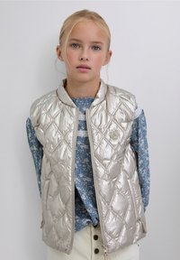 Chaleco acolchado plateado con cuello alto, cremallera frontal y bolsillos laterales, llevado sobre una blusa azul con estampado floral.