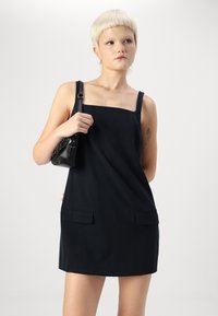 Robe noire sans manches avec décolleté carré, poches latérales et texture lisse. Un petit sac à main noir est tenu dans la main droite.