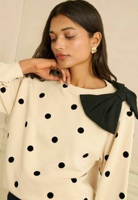 Béžový sveter s čiernymi bodkami, s veľkým čiernym mašľovým akcentom na pleci a rebrovanými manžetami. Hladká textúra látky.