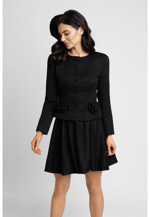 Kobieta ubrana w czarną, teksturowaną kurtkę zapinaną na guziki z kwiatowymi detalami w talii oraz dopasowaną rozkloszowaną spódnicę, pozująca na jednolitym tle.