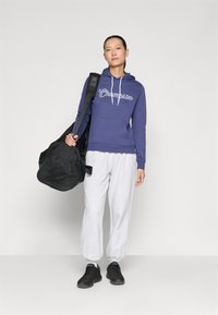 Purple hoodie with white "Champion" logo, grey sweatpants, and black shoes. Carrying a black duffel bag with mesh detailing.
