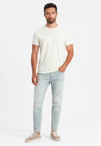 Light cream crew-neck t-shirt with a subtle pattern, paired with light blue distressed jeans and taupe sneakers, featuring white soles.