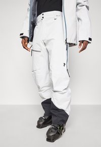Pantalones de esquí impermeables blancos con acentos negros, bolsillos con cremalleras y un ajuste relajado. Combinados con botas de esquí negras y una chaqueta en capas.