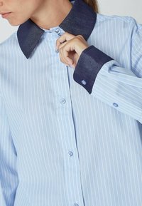 Cămașă cu dungi bleu deschis, cu guler și mansete din denim închis. Prezintă închideri cu nasturi și o croială lejeră. Dungile sunt subtile și uniform distribuite.