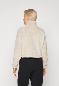 Veste en faux fur beige avec un col haut, un design court et des manches longues. Matériau texturé et doux. Portée avec un pantalon sombre.