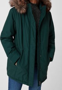 Parka matelassée verte avec capuche doublée de fourrure, dotée d'une fermeture zippée, deux poches latérales et d'un cordon élastique à la taille. Pull noir texturé en dessous.