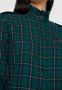 Grünes kariertes Hemd mit hohem Kragen, versehen mit schwarzen und weißen Linien. Enthält Schulterklappen und eine Brusttasche mit einem dunklen Akzent.