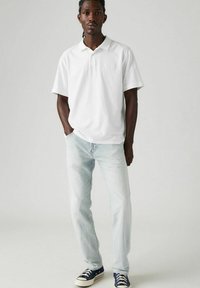White polo shirt with a simple design, paired with light blue jeans and navy sneakers. Model stands in a neutral background.