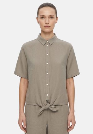 Donna che indossa una camicia taupe a maniche corte con bottoni, legata in vita, abbinata a pantaloni coordinati, in piedi davanti a uno sfondo uniforme.