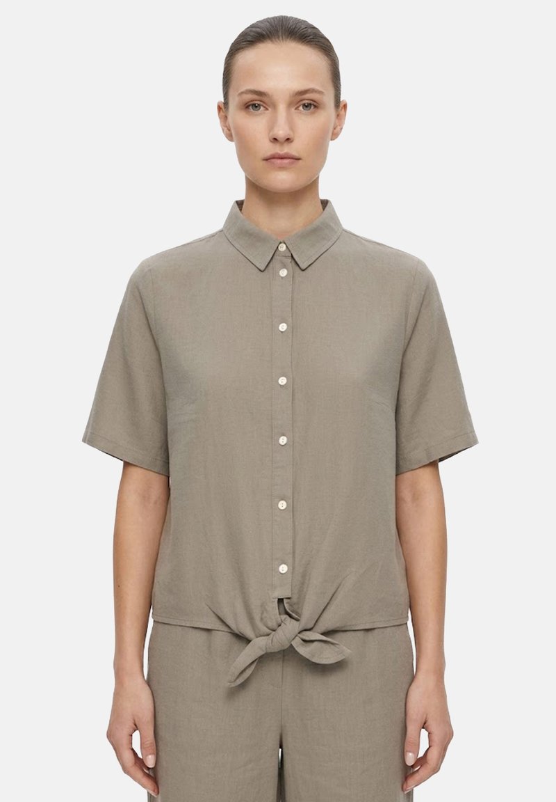 Femme portant une chemise taupe à manches courtes boutonnée et nouée à la taille, associée à un pantalon assorti, debout devant un fond uni.