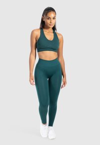 Femme debout portant un soutien-gorge de sport turquoise, un legging taille haute et des baskets blanches sur un fond gris clair uni.