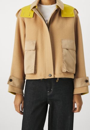 Beige uldjakke med gule skulderpaneler, store forlommer med klapper, manchet med knapper, båret over en hvid top og sorte bukser.