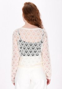 Spitzenpullover mit einer schwarzen Unterlage, der mit einem Rautenmuster, langen transparenten Ärmeln und einem gewellten Saum ausgestattet ist. Farbverlauf von Creme zu Weiß.