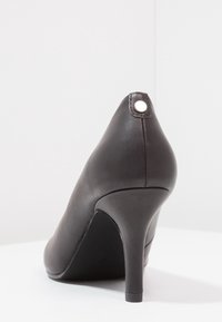 Chaussure à talon haut en cuir noir avec un bout pointu et un fin talon stiletto, présentant un petit accent circulaire à l'arrière.