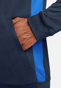 Marineblauer Sportjacke mit blauen Netz-Seitenteilen; zeigt eine Hand in der Seitentasche, die einen strukturierten Stoff und einen gerippten Bund präsentiert.