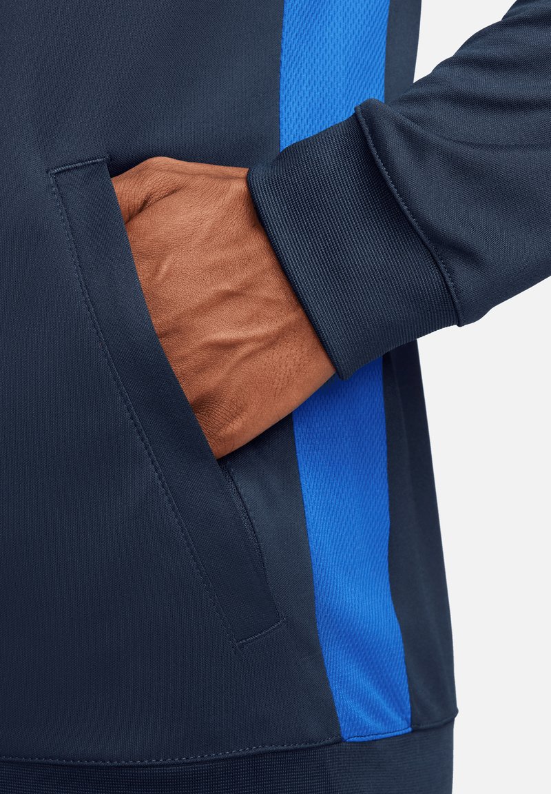 Marineblauer Sportjacke mit blauen Netz-Seitenteilen; zeigt eine Hand in der Seitentasche, die einen strukturierten Stoff und einen gerippten Bund präsentiert.