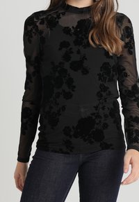 Svart genomskinlig långärmad topp med blommönster i sammet, hög krage och figurnära design, tillsammans med mörkblå jeans.