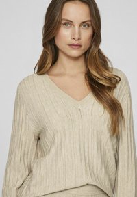 V-Ausschnitt gestrickter Pullover in hellbeige, mit vertikalem Rippmuster und oversized Passform. Weicher Stoff mit subtilen Details.