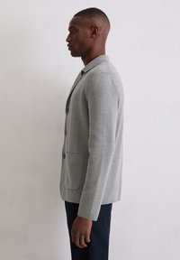 Grau gestrickter Blazer mit langen Ärmeln, Reverskragen, einer Knopfverschluss und Seitentasche, getragen über dunklen Hosen. Glatte Textur, taillierte Passform.