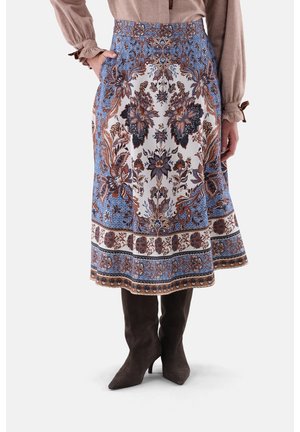 Frau trägt einen blau-braun geblümten Midirock, eine beige langärmelige Bluse und dunkelbraune Stiefel mit Absatz, die Hände in den Taschen.
