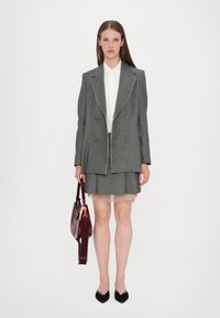 Blazer gris à double boutonnage avec garniture blanche, associé à une jupe assortie. Couche plissée transparente en dessous, sac à main bordeaux et escarpins noirs.