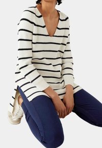 Suéter de punto a rayas en blanco y azul marino, con escote en V, mangas largas, aberturas laterales y un ajuste relajado, combinado con pantalones azul marino y zapatos blancos.