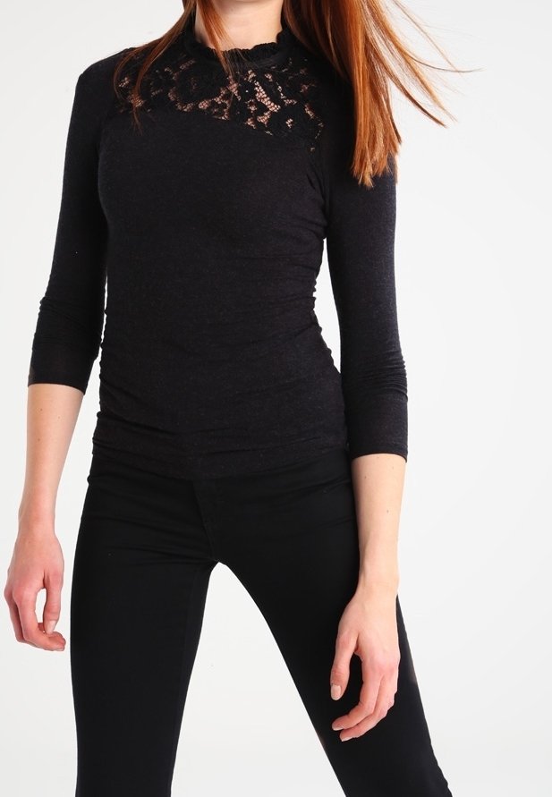 Femme portant un haut noir ajusté avec un détail en dentelle au niveau de l'encolure et des manches trois-quarts, associé à un pantalon noir sur fond blanc.