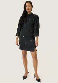 Robe noire avec des embellissements blancs décoratifs, devant boutonné, manches bouffantes et fente sur le côté. Tissu texturé et coupe ajustée.
