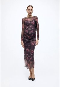 Διάφανο, μακρυμάνικο φόρεμα με λουλουδάτο μοτίβο σε σκούρο μοβ, με πιέτες, μήκος μέχρι τον αστράγαλο, συνδυασμένο με μαύρα τακούνια και μικρό μαύρο τσαντάκι.