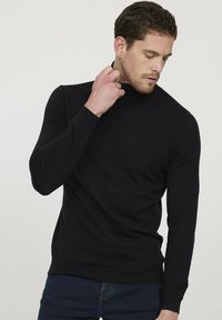 Pull à col roulé noir en tissu doux, avec des manches longues et une coupe ajustée, associé à un jean bleu.