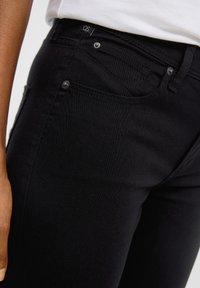 Jean stretch noir avec une coupe ajustée, poches avant et fermeture à bouton. Le haut présente une étiquette QS, avec des détails de couture subtils.