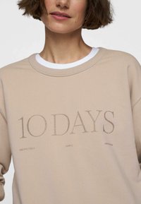Beige sweatshirt met de tekst "10 DAYS" in een glanzend lettertype. Crew neck ontwerp met een witte binnenkraag en eenvoudige gedrukte details eronder.