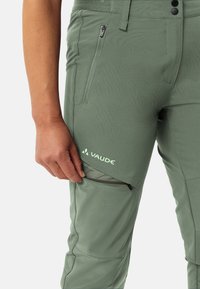 Groene wandelpantalon met een ritszak, mesh-details en een flexibele pasvorm. Branding is zichtbaar op het onderbeen.