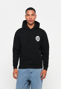 Črni kapuco s prednjim žepom, ki ima krožni beli logotip na levem prsnem delu. Material se zdi mehak in podložen s flisom.