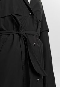 Svart trenchcoat med bälte i midjan, knappdekorationer och ett lätt tyg med en slät yta. Faller till höfthöjd med en lagerdesign.