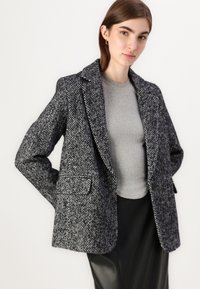 Blazer en chevrons gris avec de grands revers, deux poches avant et une coupe décontractée, porté sur un haut en maille gris et une jupe en cuir noir.