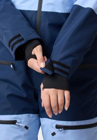 Navy- und hellblaue wasserdichte Jacke mit schwarzen Akzenten, verstellbaren Manschetten und Reißverschlüssen an den Taschen. Verfügt über Klettverschluss an den Ärmeln.